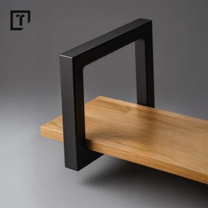Könnte beinhalten: Ein rechteckiges schwebendes Regal mit einer natürlichen Holzoberfläche und einem schwarzen Metallrahmen. Das Regal besteht aus massivem Holz. Der schwarze Metallrahmen bildet eine quadratische Form um das Regal und bietet Halt und eine moderne Ästhetik. Die Länge beträgt ca. 60 cm.