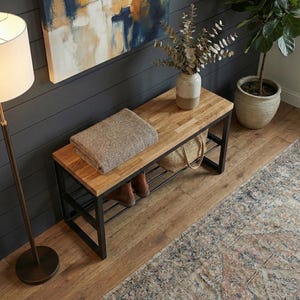 May include: A wooden bench with a black metal frame, featuring a light brown top and a lower shelf. A folded brown and beige blanket rests on the bench. A vase with dried branches and a woven bag are also visible. The bench is placed in a room with hardwood flooring.