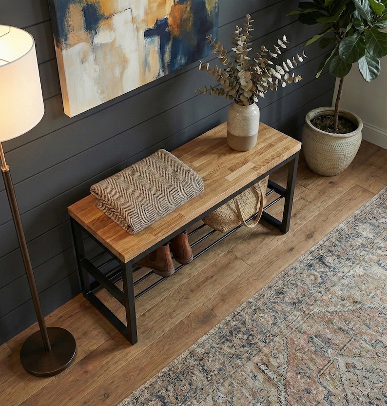 May include: A wooden bench with a black metal frame, featuring a light brown top and a lower shelf. A folded brown and beige blanket rests on the bench. A vase with dried branches and a woven bag are also visible. The bench is placed in a room with hardwood flooring.