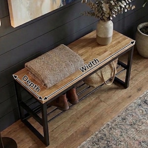 May include: A wooden bench with a black metal frame. The bench has a light brown top and a lower shelf. A folded brown and beige blanket rests on the bench. The bench is 122 cm wide and 41 cm deep.