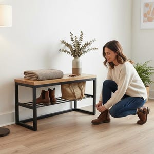 May include: A wooden and black metal shoe bench with a folded blanket, a vase with greenery, and a woven tote bag. Brown leather boots are on the lower shelf. A woman is tying her brown leather boots. A floor lamp is to the left.