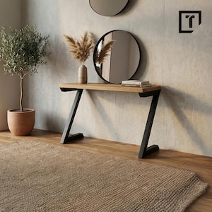Op de afbeelding: Een moderne console met een licht houten blad en zwarte metalen Z-vormige poten. Boven de tafel hangt een ronde zwarte spiegel en er staat een vaas met gedroogde bloemen op. Links staat een kleine boom in een pot.
