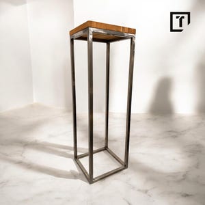Könnte beinhalten: Ein hoher, rechteckiger Metallständer mit einer Holzplatte. Der Ständer hat ein schlichtes, modernes Design und ist aus gebürstetem Metall gefertigt.