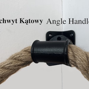 Può includere: Maniglia ad angolo in metallo nero con una corda naturale attaccata. La maniglia è montata su una parete bianca. Il testo "Uchwyt Katowy Angle Handle" è visibile sopra la maniglia.