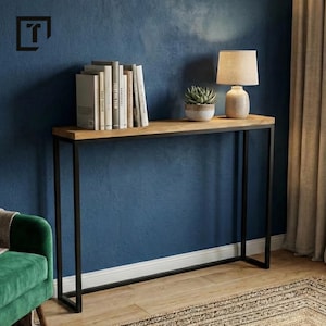 Könnte beinhalten: Eine moderne Konsolentisch mit einer hellen Holzplatte und einem schwarzen Metallrahmen. Der Tisch steht vor einer blauen Wand und trägt Bücher, eine kleine Lampe und eine Topf-Sukkulente. Der Raum verfügt außerdem über einen grünen Samtsessel und einen gemusterten Teppich.