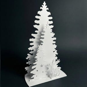 Könnte beinhalten: Weiße Papier-Weihnachtsbaumdekoration mit mehrschichtigem Design. Der Baum hat ausgeschnittene Sterne und eine Rentiersilhouette am Fuß, vor dunklem Hintergrund. Diese festliche Dekoration ist ideal für Feiertagsausstellungen.