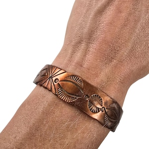 May include: A copper-colored cuff bracelet with intricate stamped designs. The bracelet features a repeating pattern of stylized sunbursts, teardrops, and circles. The metal has a warm, aged patina, suggesting it is vintage or handcrafted.