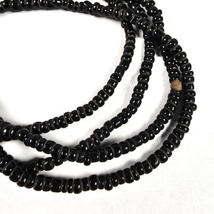May include: Close-up of several strands of black beaded necklaces. The beads are small, cylindrical, and tightly strung together, creating a smooth, glossy surface. The necklaces are arranged in a layered, intertwined pattern against a white background.