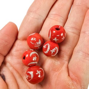 May include: Five red ceramic beads with white painted designs are held in a hand. The beads are round with a hole through the center. Each bead has a unique white design, including crescent moons and stars.