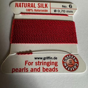 May include: Red natural silk thread for stringing pearls and beads. The spool is labeled "NATURAL SILK 100% Naturseide No. 6 Ø 0,70 mm" and "www.griffin.de For stringing pearls and beads GRIFFIN since 1866 Germany".