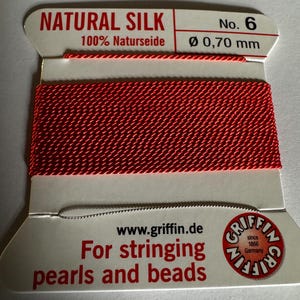 May include: Red natural silk thread for stringing pearls and beads. The spool is labeled "NATURAL SILK 100% Naturseide No. 6 Ø 0,70 mm" and "www.griffin.de For stringing pearls and beads" with the Griffin logo.