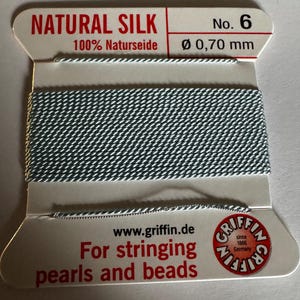 May include: A spool of light blue natural silk thread for stringing pearls and beads. The spool is labeled "NATURAL SILK 100% Naturseide No. 6 Ø 0,70 mm" and "www.griffin.de For stringing pearls and beads GRIFFIN since 1866 Germany".