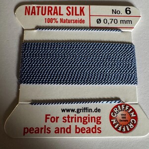 May include: A spool of natural silk thread for stringing pearls and beads. The thread is blue with a white stripe and is labeled "Natural Silk 100% Naturseide No. 6 Ø 0,70 mm". The label also includes the website www.griffin.de and the Griffin logo.