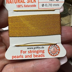 May include: A spool of gold-colored natural silk thread for stringing pearls and beads. The label reads "NATURAL SILK 100% Naturseide No. 6 Ø 0,70 mm" and "www.griffin.de For stringing pearls and beads since 1865 Germany".