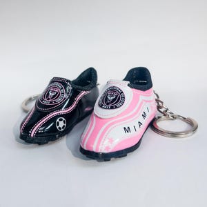 May include: Miniature soccer cleats keychains, one black and one pink, with the "Internacional de Futbol Miami" logo. The black cleat has a soccer ball graphic. Both have silver key rings.