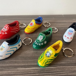 May include: A collection of miniature soccer shoe keychains in various colors, each representing a different country. The keychains feature the flags and names of countries like Canada, Argentina, Mexico, USA, and Brazil. The shoes are made of a glossy material.