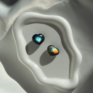 Può includere: Due pietre di labradorite a forma di cuore con riflessi iridescenti blu, verdi e oro poggiano su un piatto di ceramica bianco dalla forma organica. Le pietre sono su una superficie bianca strutturata.