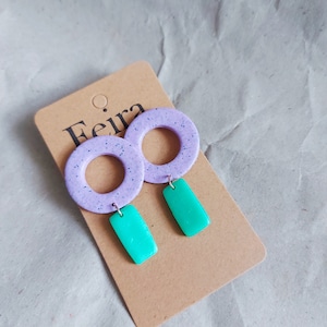May include: A pair of lavender and teal earrings with a circle and rectangle design. The earrings are on a brown card with the word "Feira" printed on it.
