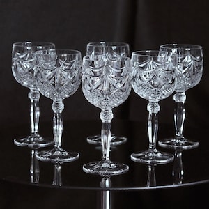 May include: Six clear crystal wine glasses with detailed cut patterns. The glasses feature a classic design with a rounded bowl, a slender stem, and a circular base. The glasses are arranged on a reflective surface against a dark background.
