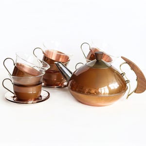 May include: A copper tea set with a teapot, cups, and saucers. The teapot has a wooden handle and lid. The cups are clear glass with copper bottoms and handles. The set is on a white background.