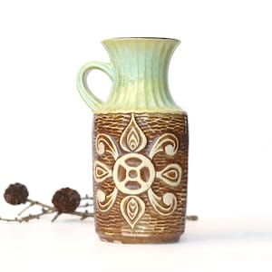 Könnte beinhalten: Eine Vintage-Keramikvase mit Henkel. Die Vase hat einen braunen, strukturierten Körper mit einem cremefarbenen Blumendesign. Der obere Teil ist hellgrün mit vertikalen Rillen. Die Vase ist ein dekoratives Stück.