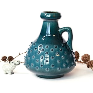 Könnte beinhalten: Eine tealblaue Keramikvase mit einer strukturierten Oberfläche und einem Henkel. Die Vase hat einen runden Körper und einen schmalen Hals.