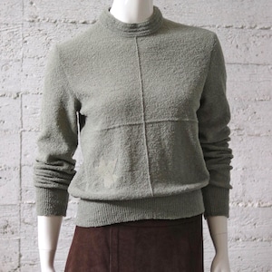 Könnte beinhalten: Ein salbeigrüner Pullover mit Stehkragen und langen Ärmeln. Der Pullover hat eine strukturierte Optik und ein dezentes Kreuzstichmuster auf der Vorderseite. Die Bündchen und der Saum sind gerippt. Der Pullover wird auf einer Schaufensterpuppe präsentiert.