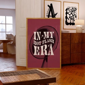 May include: A framed print with a burgundy background and the text "IN MY HOT FLASH ERA" in white. The image features a black fan design. Two other framed prints are visible in the background.
