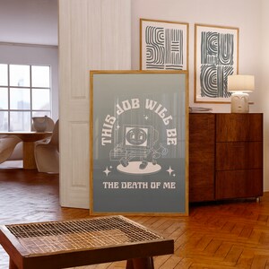 Puede incluir: Un póster enmarcado con el texto "THIS JOB WILL BE THE DEATH OF ME" y un personaje de dibujos animados de ordenador. El póster tiene un fondo degradado azul claro y gris. En el fondo se ven otras dos impresiones artísticas abstractas enmarcadas.