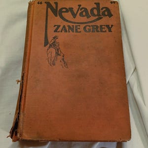 Op de afbeelding: Een oud boek met de titel "Nevada" van Zane Grey. De kaft is verbleekt oranje met zwarte letters en een illustratie van een cowboy te paard. Het boek vertoont tekenen van slijtage.