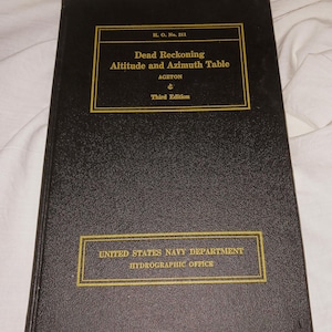 May include: A black book titled "Dead Reckoning Altitude and Azimuth Table" with gold lettering and borders. The book is a third edition by the United States Navy Department Hydrographic Office.