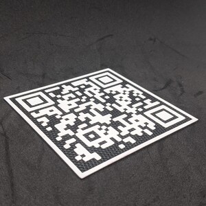 May include: A black and white QR code printed on a white square sticker. The sticker is on a black surface.