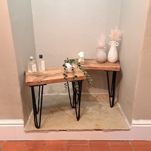 May include: Two wooden shelves with black hairpin legs, one on top of the other. The top shelf has two white ceramic vases with dried flowers. The bottom shelf has a small potted plant and two bottles of perfume.