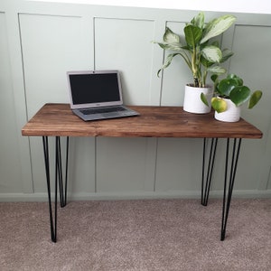 May include: A dark brown wooden desk with black hairpin legs. A laptop computer is on the desk, and two potted plants are on the right side of the desk.
