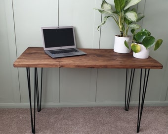 May include: A dark brown wooden desk with black hairpin legs. A laptop computer is on the desk, and two potted plants are on the right side of the desk.