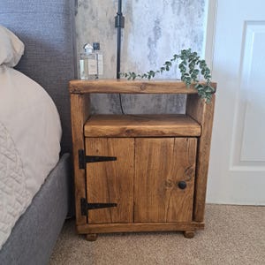 May include: A handcrafted wooden bedside table with a single door and a shelf. The table is made of light brown wood and features black hinges and a black knob. A bottle of perfume and a plant are on top.