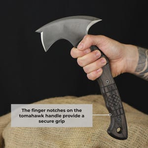 Pu&ograve; includere: Un'ascia tomahawk con un manico in legno scuro e una lama argentata curva. L'impugnatura presenta intagli per le dita per una presa sicura. L'ascia &egrave; tenuta in mano su sfondo scuro.