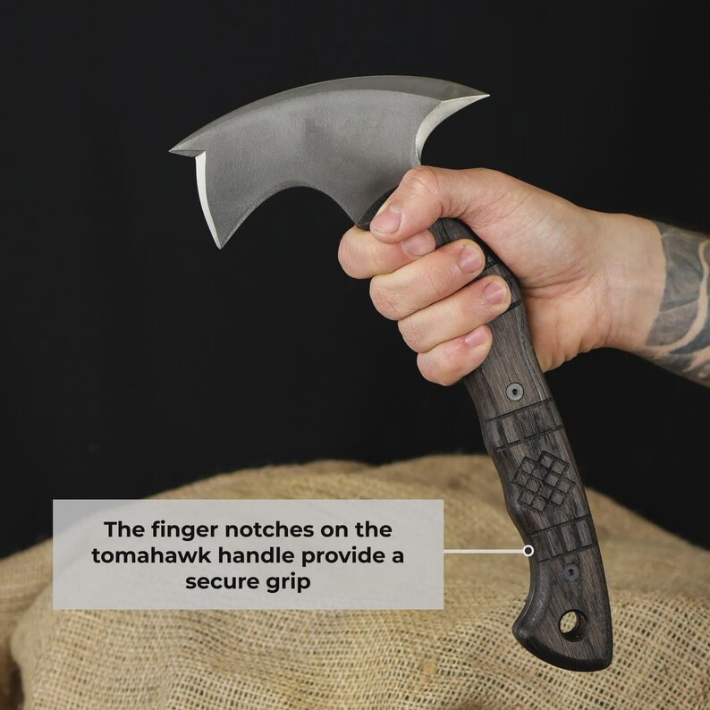 Pu&ograve; includere: Un'ascia tomahawk con un manico in legno scuro e una lama argentata curva. L'impugnatura presenta intagli per le dita per una presa sicura. L'ascia &egrave; tenuta in mano su sfondo scuro.