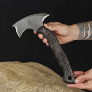 Pu&ograve; includere: Un'ascia artigianale con una lama curva color argento e un manico in legno scuro. Il manico presenta intricati disegni intagliati ed &egrave; fissato con rivetti metallici. L'ascia &egrave; tenuta su uno sfondo scuro.