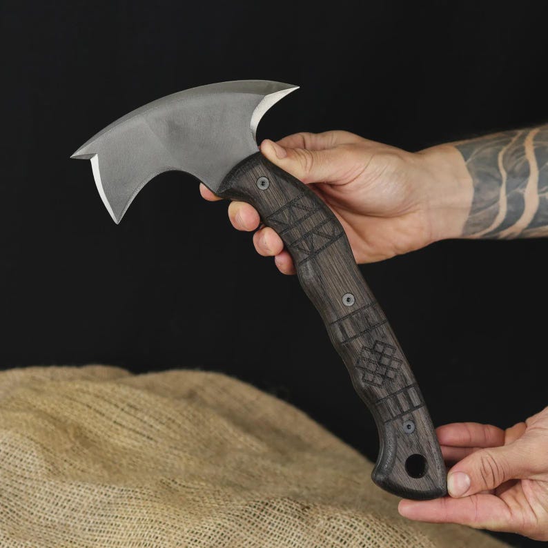 Pu&ograve; includere: Un'ascia artigianale con una lama curva color argento e un manico in legno scuro. Il manico presenta intricati disegni intagliati ed &egrave; fissato con rivetti metallici. L'ascia &egrave; tenuta su uno sfondo scuro.