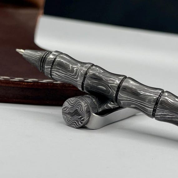 A ballpoint pen with a hand-forged Damascus steel barrel — functional desk art where every pen has a completely unique swirling pattern from the steel-folding process.