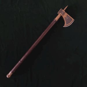 Puede incluir: Hacha medieval con un mango de madera marrón oscuro y una cabeza decorativa. La cabeza del hacha presenta filigranas doradas y una punta. La hoja tiene un borde curvo. Un arma histórica.