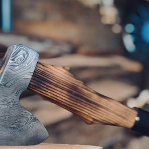 May include: Close-up of a handcrafted axe with a patterned steel blade and a wooden handle. The handle is made of light brown wood with a dark, charred finish. The axe is resting on a wooden surface, showcasing the craftsmanship.