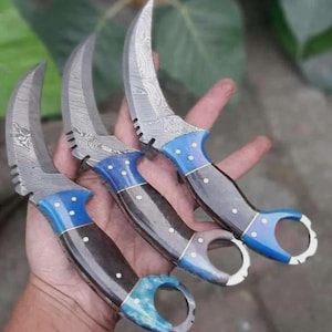 May include: Three curved karambit knives with patterned blades and blue and black handles. The blades have a textured design, and the handles feature a finger ring. The knives are held in a hand.