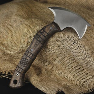 Pu&ograve; includere: Un'ascia artigianale con un manico in legno scuro con motivi geometrici incisi. La testa dell'ascia &egrave; una lama curva color argento. L'ascia &egrave; esposta su un tessuto strutturato color beige.