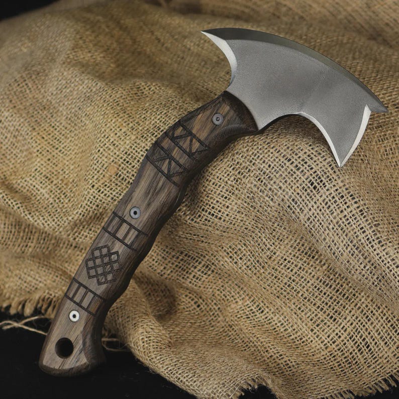 Pu&ograve; includere: Un'ascia artigianale con un manico in legno scuro con motivi geometrici incisi. La testa dell'ascia &egrave; una lama curva color argento. L'ascia &egrave; esposta su un tessuto strutturato color beige.