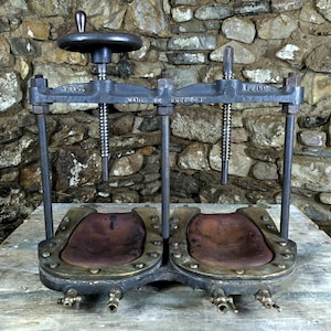 May include: Vintage cast iron press with a dark finish, featuring a large hand wheel and two pressing surfaces. The press is mounted on a wooden surface against a stone wall, showcasing its vintage design.