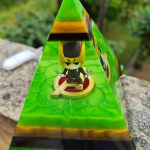 May include: A green, gold, and black pyramid-shaped resin paperweight with a small figurine of a character wearing a black suit and a gold helmet inside. The character is holding two silver daggers.