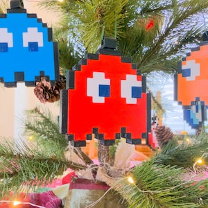 May include: Pixelated ghost ornaments in blue, red, and orange hang on a Christmas tree. Each ornament has a black outline and white eyes. The tree is decorated with pinecones and lights, creating a festive holiday scene.