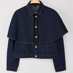 Puede incluir: Chaqueta vaquera azul oscuro con detalle de capa. La chaqueta tiene un cuello clásico, botones dorados y una silueta entallada. Cuelga de una percha de madera sobre un fondo blanco.